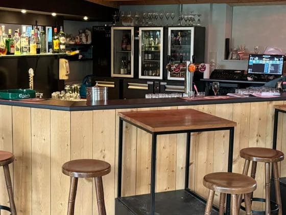 De halfronde bar in hout, met tapkraan, koelkasten en barkrukken.